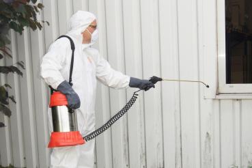 Anwendung des MESTO INOX PLUS 6 Hochdrucksprühgeräts 3595P durch eine Person in Schutzkleidung und Atemmaske bei der Außenreinigung eines Gebäudes.